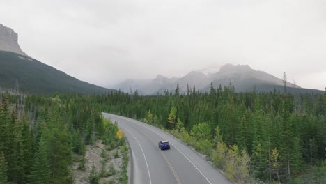 Aerial-dolly-in-of-car-driving-on-road-surrounded-by-pine-trees,-Canadian-Rockies-in-background,-Banff-National-Park,-Alberta,-Canada