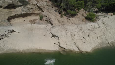 Panoramic-drone-shot-of-young-man-jumping-into-natural-water-body-from-edge-of-rock,-with-trees-in-background,-enjoying-holidays-on-solo-trip-to-El-Chorro-of-Málaga,-Spain