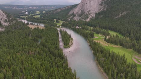 Aerial-dolly-in-over-Bow-River-between-Pine-forest-near-steep-hill,-Canadian-Rockies-in-back,-Banff-National-Park,-Alberta,-Canada