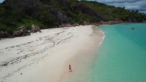 Woman-in-Red-bathing-suit-walking-down-sandy-tropical-beach-alone-on-Curieuse-Island-The-Seychelles-with-boat-anchored-off-shore