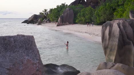 Woman-in-black-swimsuit-standing-in-water-by-Seychelles-Beach-with-boulders-at-sunset