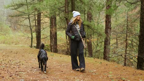 Woman-walks-in-the-green-forest-with-her-black-dog,-enjoying-and-spending-time-together-in-nature