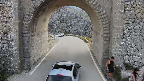Drone-following-a-car-driving-under-a-bridge-onto-a-winding-mountain-road-in-Sa-Calobra,-Mallorca,-Spain