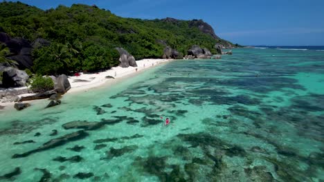 Frau-Im-Roten-Badeanzug-Paddelboot-Vor-Der-Küste-Des-Beliebten-Strandes-Der-Seychellen-Mit-Felsbrocken-Und-Weißem-Sand