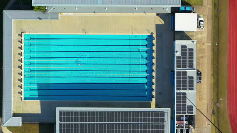 Aerial-Drone-Birds-Eye-View-Of-Blue-Olympic-Swimming-Pool-At-University,-With-Solar-Panel-Roof,-4K