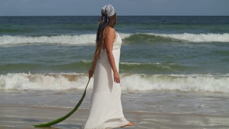 Young-girl-walking-on-the-beach-holding-a-palm-leaf-while-wearing-a-beach-dress-and-ocean-waves-crashing-the-shoreline-in-the-background