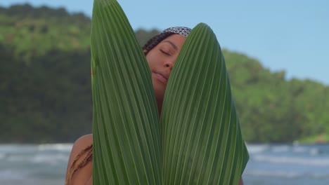 Young-lady-face-reveal-close-up-behind-a-palm-leaf-on-the-beach