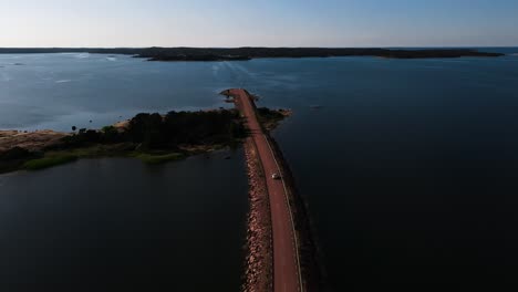 Aerial-view-following-a-car-driving-on-a-narrow-bridge-road-to-a-ferry-stop,-summer-in-Aland,-Finland