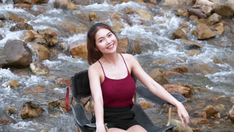 Mujer-Asiática-De-Rojo-Sentada-En-Una-Silla-Con-Grandes-Rocas-Y-Agua-Salpicando-Lentamente-Para-Relajarse-Y-Ser-Feliz-En-Las-Vacaciones-De-Verano