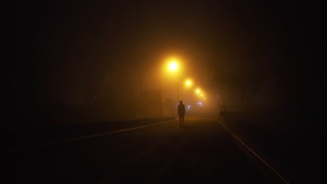 Man-in-a-hoody-walking-down-a-misty-street-at-night-with-hazy-street-lights-barely-lighting-up-the-road