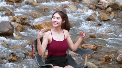 Mujer-Asiática-De-Rojo-Sentada-En-Una-Silla-Con-Grandes-Rocas-Y-Agua-Salpicando-Lentamente-Para-Relajarse-Y-Ser-Feliz-En-Las-Vacaciones-De-Verano