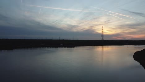 Lonely-powerline-pole-silhouette-standing-behind-river-Scheldt,-aerial-fly-towards-view
