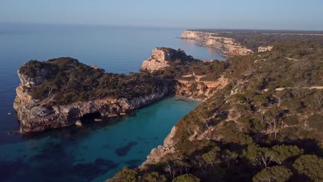 Aerial-drone-flight-showing-beautiful-Calo-des-Moro-Bay-with-rocky-coastline-lighting-by-sunset-on-Mallorca-Island---Mediterranean-Sea-in-background-in-the-evening