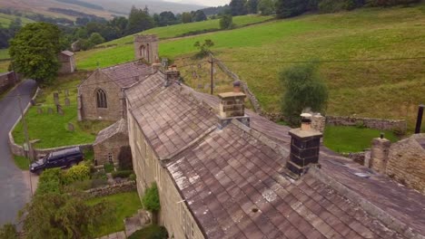 Drone-aerial-shot-of-old-stone-cottage-chimney-smoke-in-Yorkshire-village-Churchyard-comes-into-view