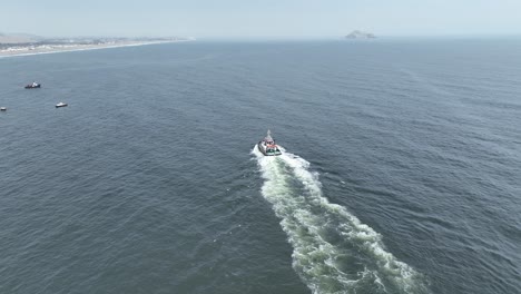 Statische-Luftaufnahme-Mit-Fokus-Auf-Einem-Sich-Bewegenden-Boot-Im-Pazifischen-Ozean-Während-Ruhiger-Wellen-Im-Meer-Mit-Blick-Auf-Andere-Boote-Und-Den-Strand-Von-Pucusuna-Beach-In-Peru
