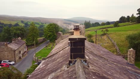 Drone-aerial-reveal-shot-of-old-stone-cottage-roof-with-chimney-smoke-over-Yorkshire-village