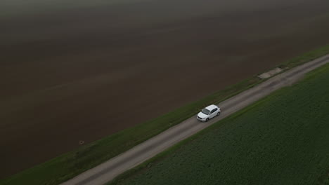 White-car-driving-country-road-between-fields,-following-revealing-drone-view,-bad-cloudy-weather-and-fog