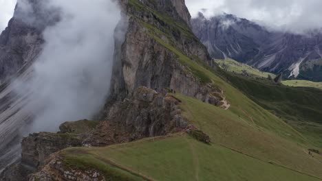 Aerial-drone-flight-showing-couple-on-green-edge-of-mountain-watching-majestic-summit-of-Dolomites-Mountains,Italy