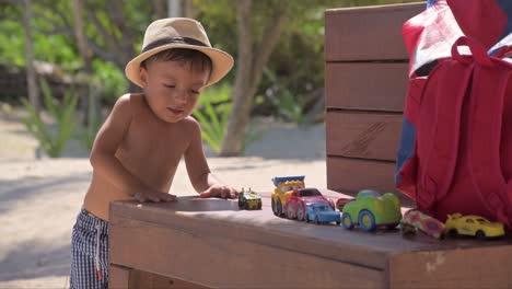 Young-latin-baby-boy-playing-with-his-car-toys-on-a-bench-covered-in-sand-at-the-beach-on-a-sunny-summer-day-wearing-a-hat