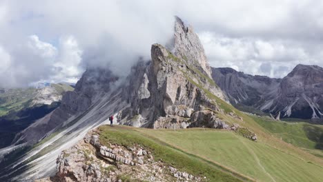Aerial-view-of-hiker-on-green-mountaintop-of-Seceda-Ridgeline-Hike-watching-Dolomites-mountains-during-cloudy-day,Italy