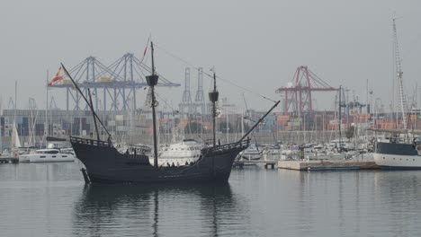 Ferdinand-Magellan-Nao-Victoria-carrack-boat-replica-with-spanish-flag-enters-wharf-in-Valencia-with-containers-and-cranes-in-the-background-in-slow-motion-60fps