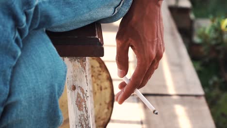 Slow-motion-video---Close-up-of-hands-of-a-young-man-holding-a-cigarette-while-sitting-relaxed-on-the-wooden-chairs-and-wearing-jeans
