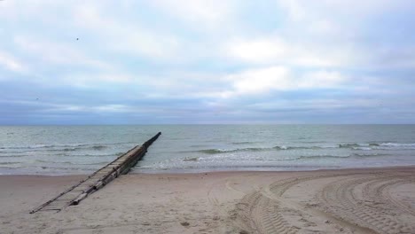Establishing-aerial-view-of-Baltic-sea-coast-on-a-overcast-day,-old-wooden-pier,-white-sand-beach,-low-waves-crushing-against-the-coast,-climate-changes,-low-wide-angle-drone-shot-moving-forward
