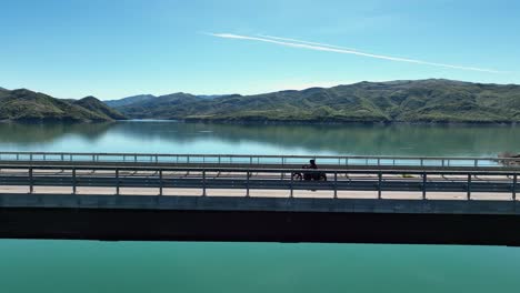 Biker-crossing-a-bridge-overlooking-a-beautiful-blue-lake-areal-shot