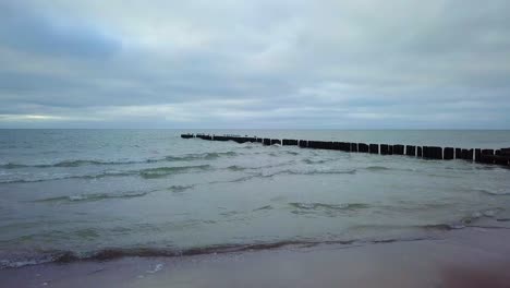 Establishing-aerial-view-of-Baltic-sea-coast-on-a-overcast-day,-old-wooden-pier,-white-sand-beach,-low-waves-crushing-against-the-coast,-climate-changes,-low-wide-angle-drone-shot-moving-forward