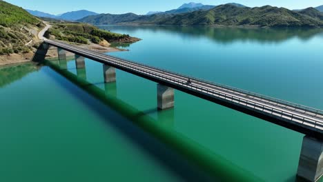 Areal-shot-of-motorcycles-crossing-a-long-empty-bridge-over-a-calm-blue-lake-near-the-hills