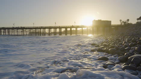 Dolly-shot-moving-back-of-the-Ventura-Beach-with-waves-crashing-along-the-rocky-shores-with-the-Ventura-Pier-in-the-background-at-sunset-located-in-Southern-California