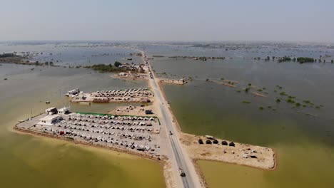 Aerial-View-Of-Lone-Elevated-Road-With-Land-Housing-Makeshift-Camps-For-Flood-Refugees-Surrounded-By-Expansive-Flood-Waters-In-Rural-Jacobabad,-Sindh