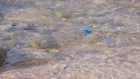 Plastic-blue-bottle-cap-floating-in-water-being-take-by-current,-slowmotion-pan