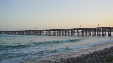 Panning-shot-of-Ventura-Pier-with-waves-of-the-Pacific-Ocean-rolling-to-the-shore-with-a-bright-orange-sunset-reflecting-on-the-water-located-in-Southern-California