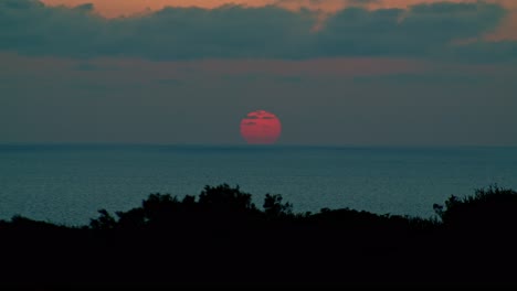 Astonishing-red-sun-setting-below-sea-horizon,-cumulus-grey-clouds,-static