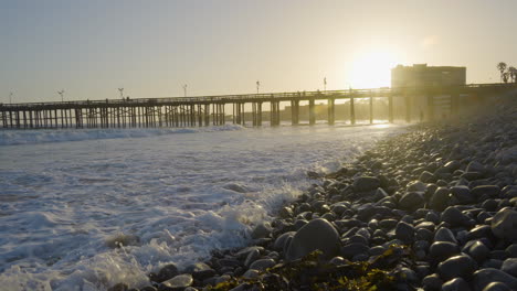 Dolly-shot-moving-forward-along-the-rocky-shore-of-a-Southern-California-Beach-while-waves-are-crashing-in-with-Venture-Pier-in-the-background-located-in-Southern-California