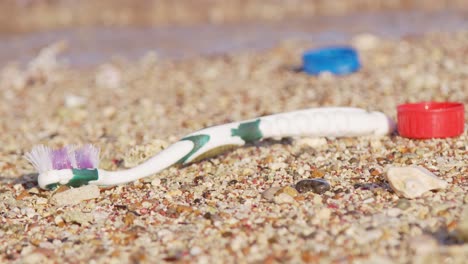 Plastic-old-toothbrush-and-bottle-caps-washed-up-on-sandy-beach-being-washed-away-by-current-and-waves