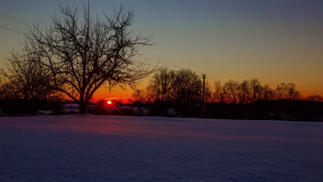 Brilliant-orange-sunset-on-a-winter-day-over-European-snowy-countryside---time-lapse
