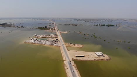 Vista-De-Drones-Del-Campamento-Improvisado-Con-Tiendas-De-Campaña-Para-Albergar-A-Los-Refugiados-De-Las-Inundaciones-Al-Lado-De-Una-Carretera-Elevada-Rodeada-De-Amplias-Aguas-De-Inundación-En-La-Zona-Rural-De-Jacobabad,-Sindh