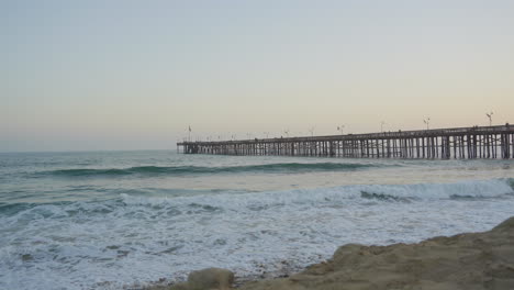 slow-motion-Panning-shot-of-waves-rolling-through-the-Pacific-Ocean-crashing-into-the-shores-of-Ventura-Beach-with-the-pier-and-sunset-in-the-background-located-in-Southern-California