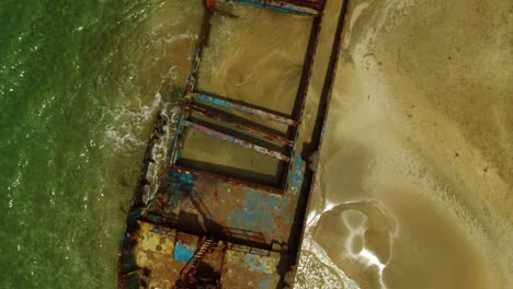 Small-waves-wash-across-a-rusted-abandoned-shipwreck-on-a-golden-sand-beach,-Manzanillo,-aerial-top-down