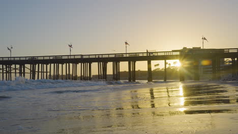Dolly-shot-moving-backwards-along-the-the-Ventura-Beach-while-waves-crash-along-the-shore-with-the-Ventura-Pier-and-sunsetting-in-the-background-located-in-Southern-California