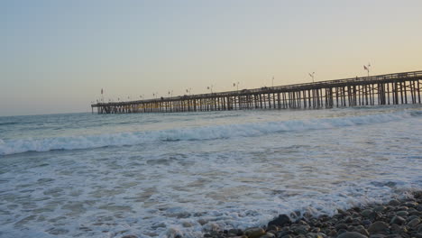 Panning-shot-of-waves-crashing-along-the-shores-of-Ventura-Beach-with-Pier-and-sunsetting-in-the-background-located-in-Southern-California