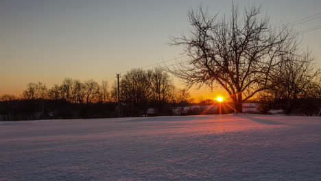An-orange,-glowing-sunrise-over-a-winter,-countryside-meadow-covered-in-fresh-snow---time-lapse