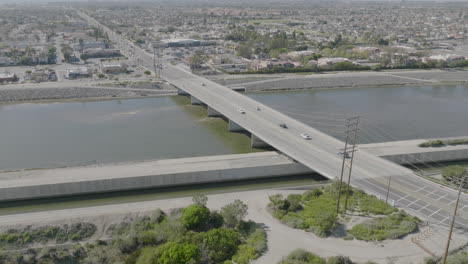 Flyover-aerial-shot-of-cars-going-over-a-bridge-over-a-river-in-Costa-Mesa,-California