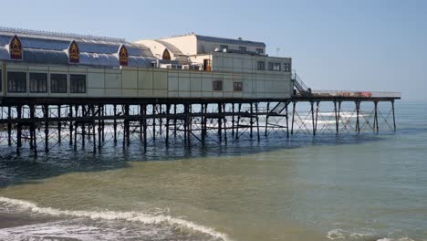 Waves-wash-on-the-beach-underneath-the-pier-in-Aberystwyth,-Ceredigion,-Mid-Wales-on-a-clear-day-with-bright-blue-sky