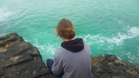 Back-View-Of-A-Sad-Male-Teenager-Sitting-On-Cliff-Edge-Looking-Down-At-Ocean-Waves-Hitting-The-Rock---High-Angle
