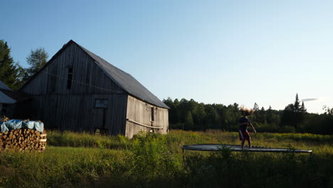 Kid-using-a-trampoline-at-a-farm-with-wood-stack,-barn,-forest-and-field