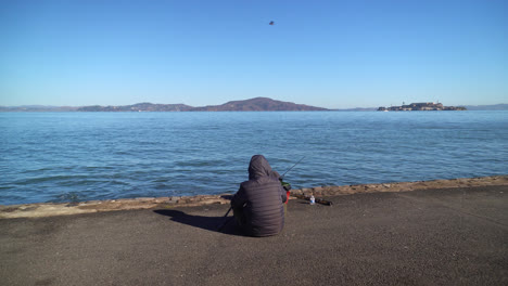 Person-Fishing-And-Sitting-On-Seaside-With-Alcatraz-Island-In-The-Background-In-San-Francisco-Bay