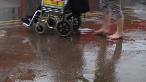 Eine-Barfüßige-Frau-Läuft-Im-Regen-Durch-Pfützen,-Während-Sie-Einen-Rollstuhl-Schiebt